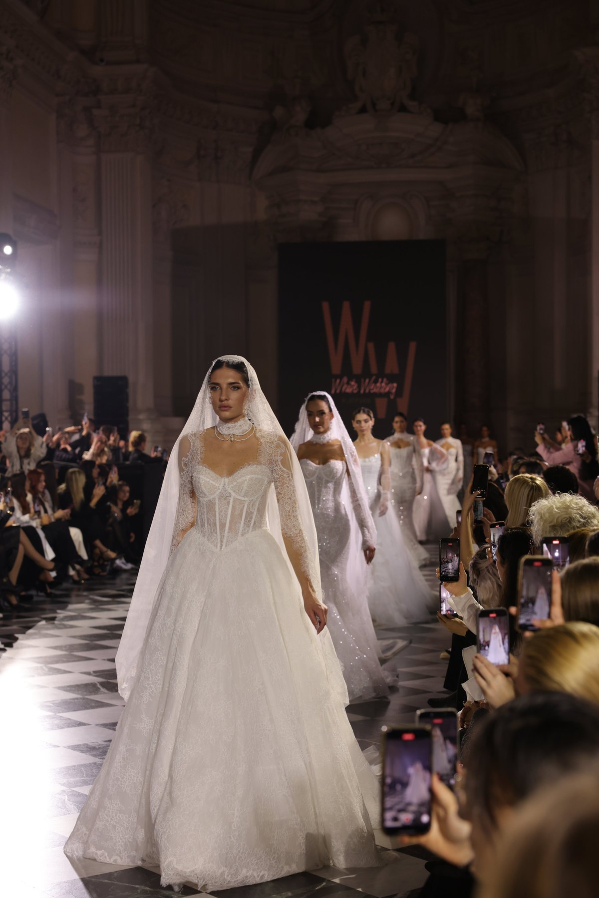 Bride in white wedding gown and veil walking down runway at fashion show, audience members watching and taking photos