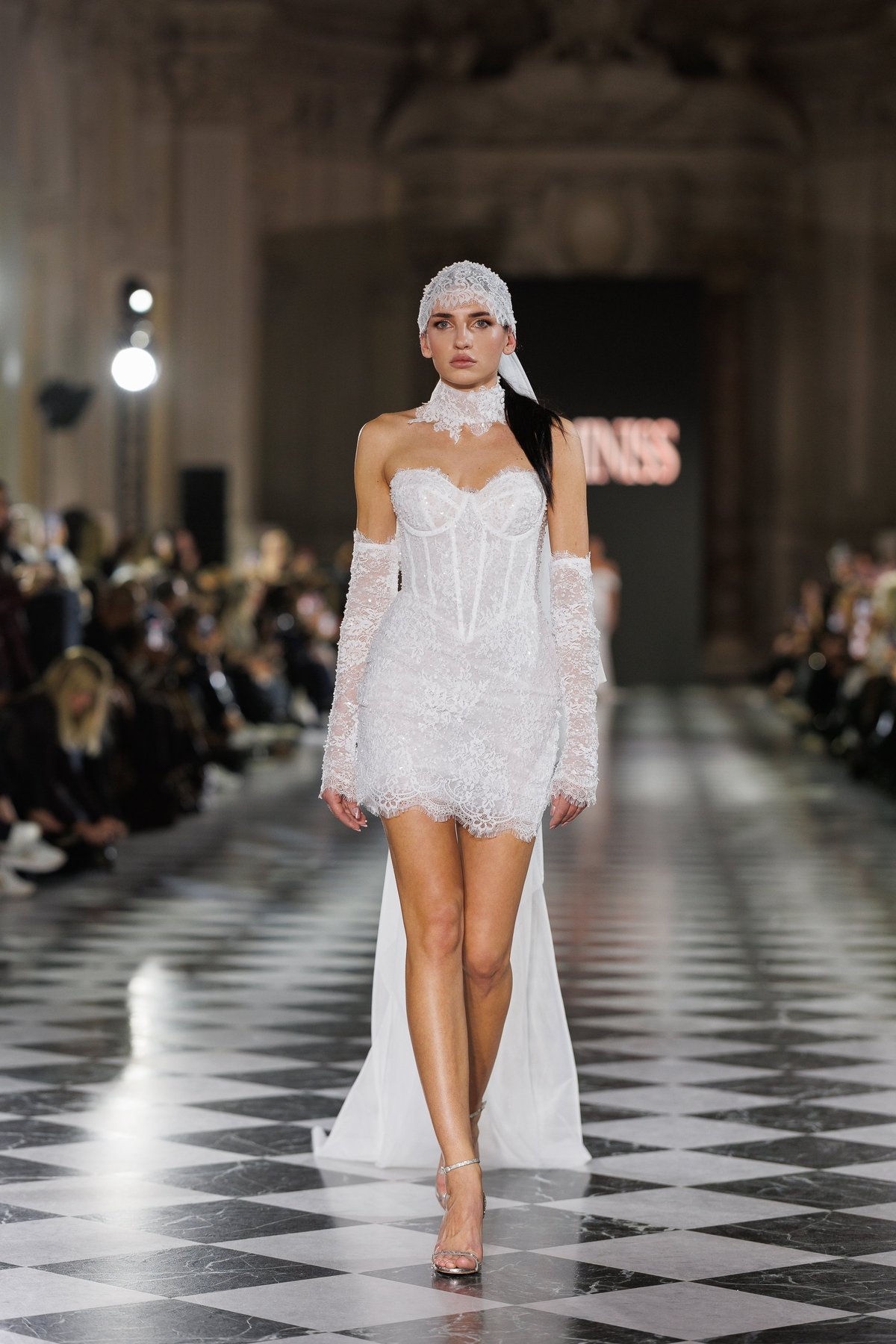 Model walking down a fashion runway in a white lace strapless dress with matching head wrap and long gloves
