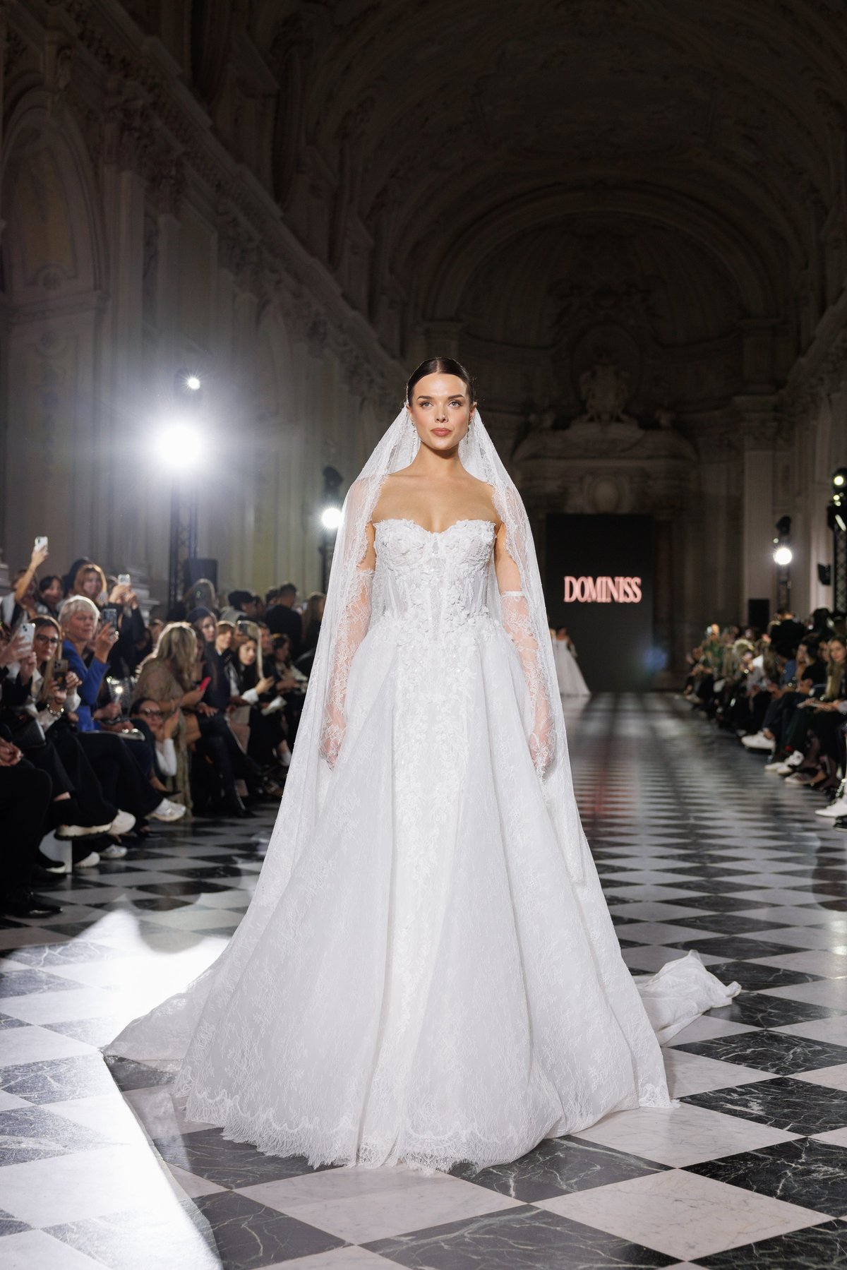Model wearing an elegant white lace wedding gown with veil on a cathedral runway during a bridal fashion show