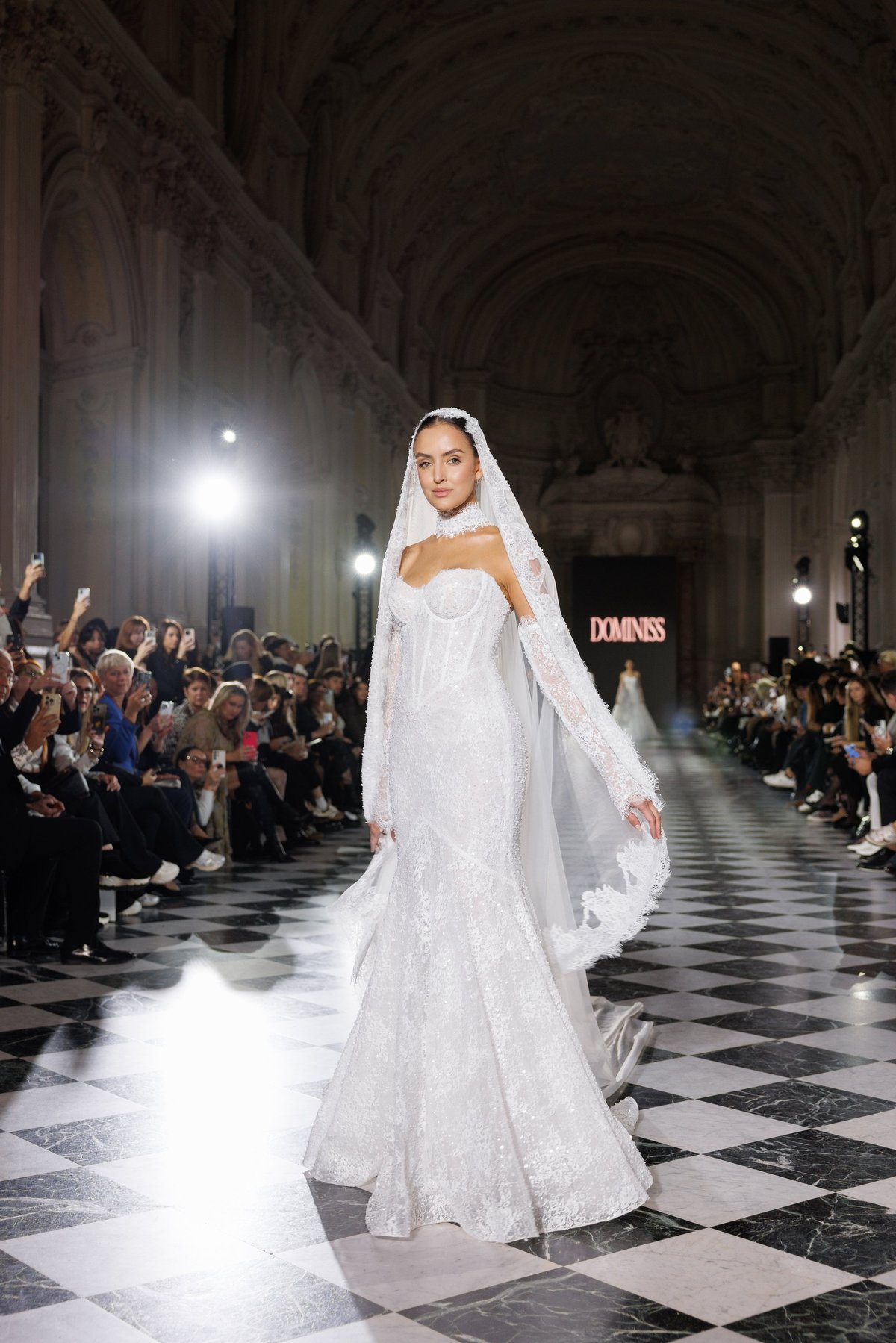 Model wearing an ornate white wedding gown with veil walks down a cathedral runway during a fashion show