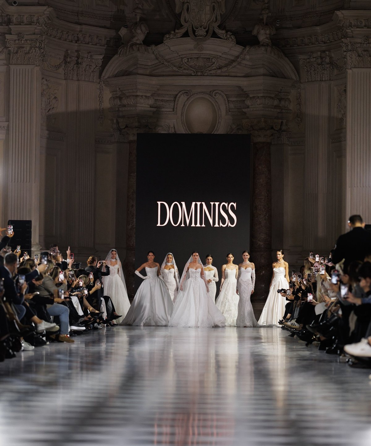 Fashion models wearing white gowns walk down a runway during a Dominiss fashion show in an elegant venue with ornate columns.