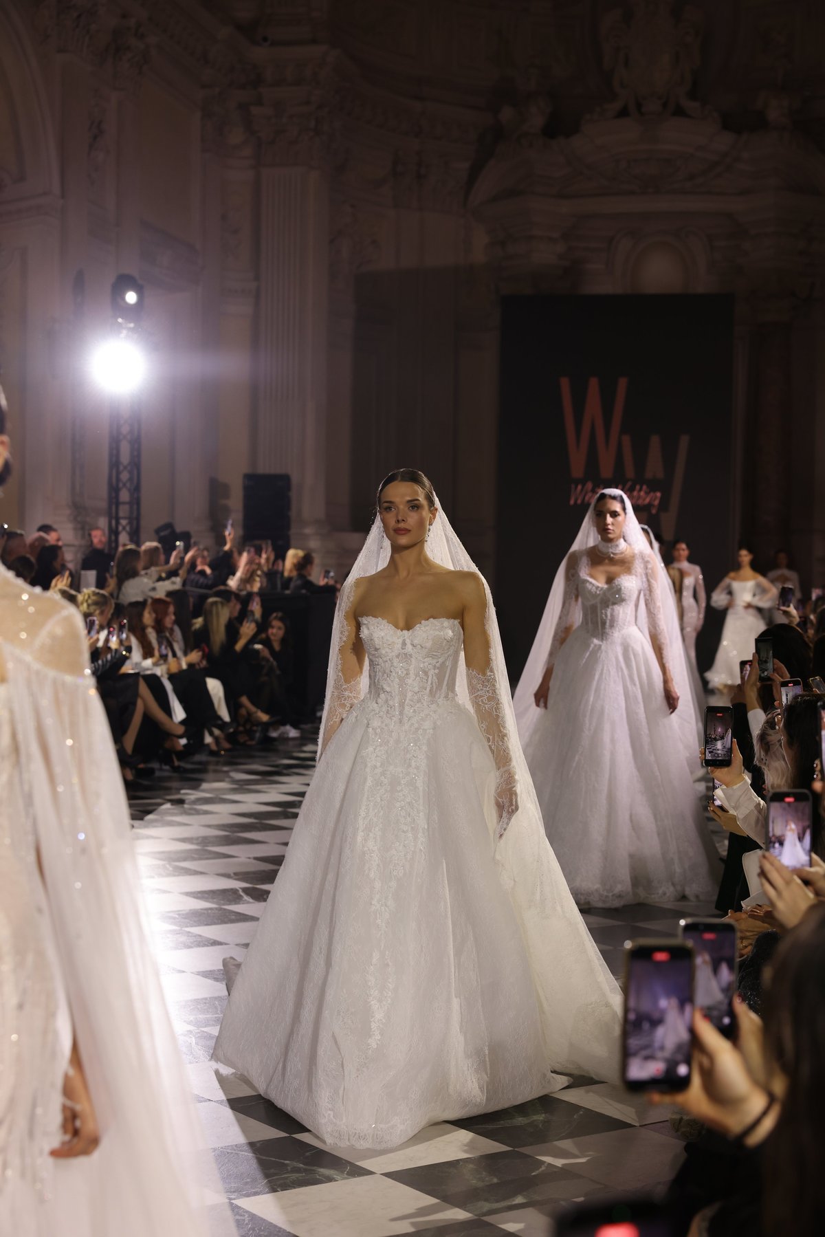 Model wearing an ornate beaded white bridal gown with veil walking on runway during formal wedding dress fashion show