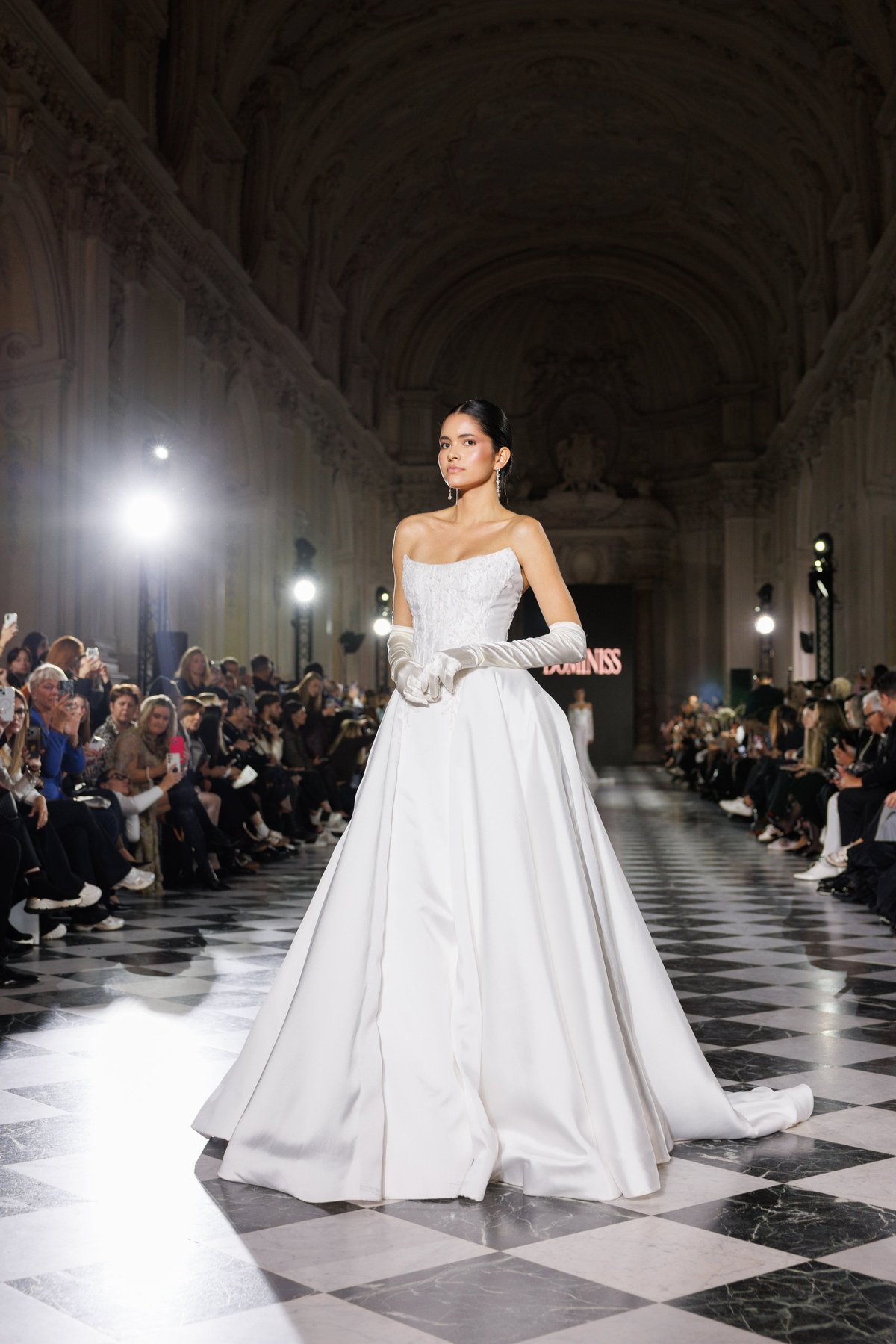 Model wearing elegant white strapless gown on runway in grand arched hallway with audience seated along sides