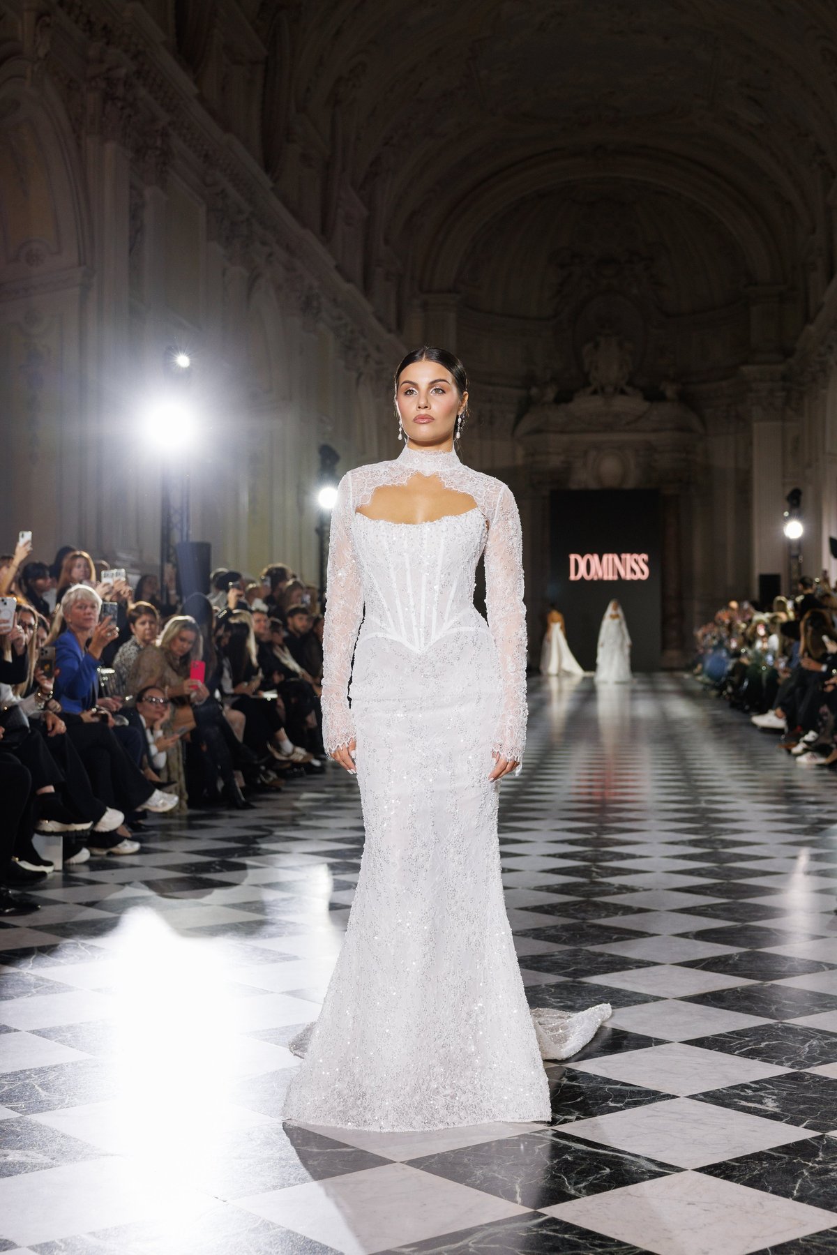 Model wearing white lace gown on runway in ornate cathedral setting with audience seated along sides