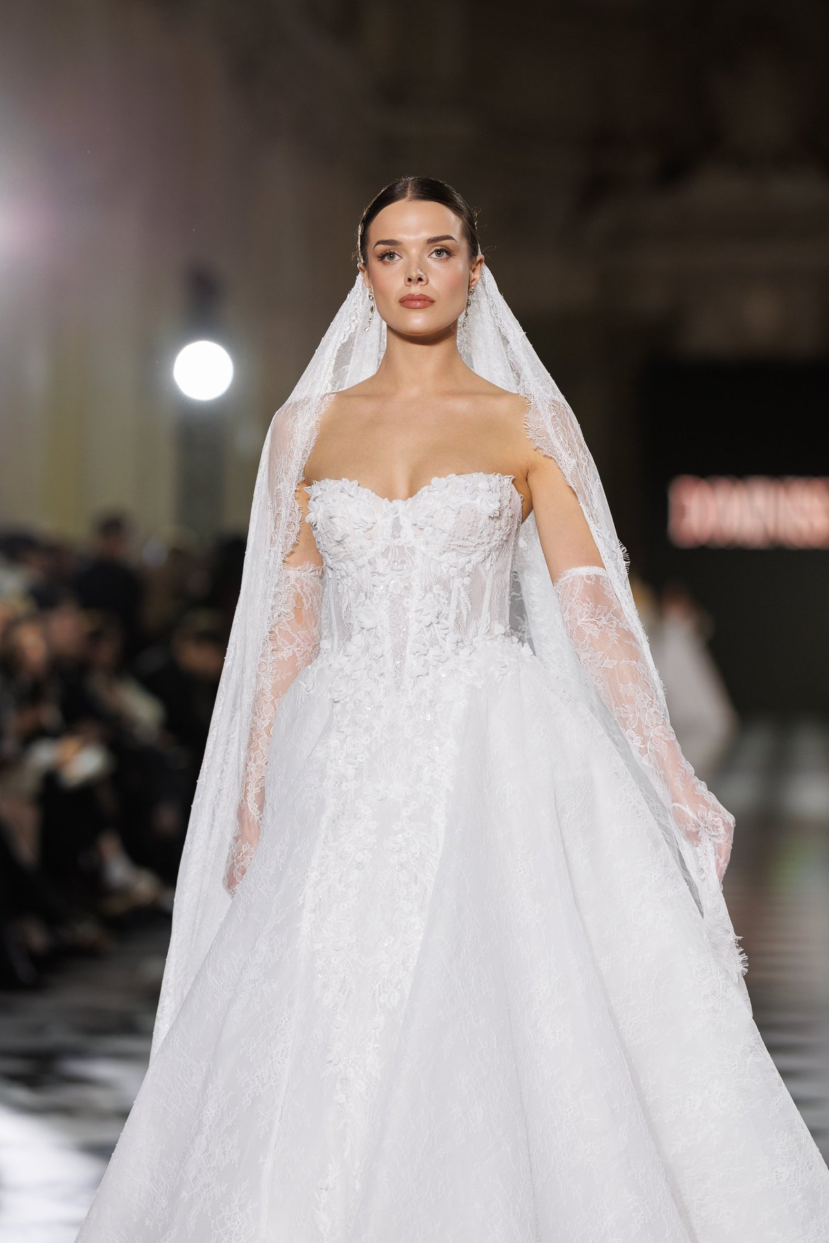 Model wearing an ornate white wedding gown with sweetheart neckline and bridal veil on a fashion runway